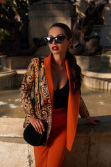 Fashion style outdoor portrait. Elegant woman in colorful orange suit with black handbag standing and posing against fountain in old town on a sunny evening, Sunny evening. Tropical city