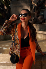 Fashion style outdoor portrait. Elegant woman in colorful orange suit with black handbag standing and posing against fountain in old town on a sunny evening, Sunny evening. Tropical city