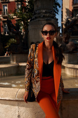Fashion style outdoor portrait. Elegant woman in colorful orange suit with black handbag standing and posing against fountain in old town on a sunny evening, Sunny evening. Tropical city