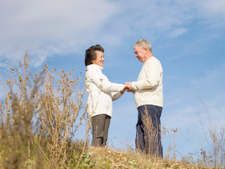 Elderly happy couple in white knitted sweaters are standing holding hands on a blue naba background in autumn afternoon. Walk the elderly. Love