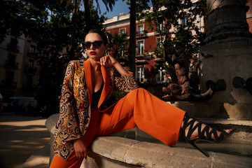 Fashion style outdoor portrait. Elegant woman in colorful orange costume, sunglasses and black shoes sitting on stone fence of fountain on a sunny evening in tropical city