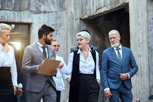 Group Of Businessmen In Offic Wear Move In Street. Blond Woman Point Out Papier Where Man Take Notes. Other People Follow