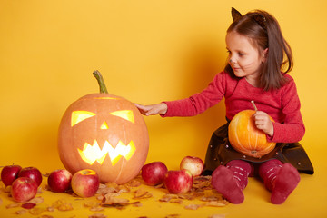 Fototapeta premium Indoor shot of cute dark haired girl sitting on floor over backgroundof autumn scenery in studio. Autumn harvest - pumpkin, apples. Little kid playing with Jack o'Lantern. Halooween concept.