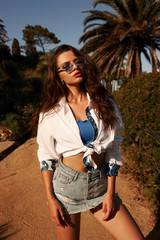 Beautiful stylish woman walking in tropical park with palms on a sunny evening. Tanned girl in jeans skirt, blue top, white blouse and sunglasses. Relaxing vacation concept