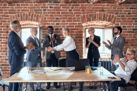Group of businessmen congratulate colleague with good decision in office. Adult caucasian man wear blue suit handshake aduld blond woman dress white shirt. She smile, look at him. Others clap, smile