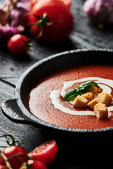 Tomato soup with cream, croutons and basil in a black bowl surrounded with tomatoes and garlic on black wooden background. Copy space for text. Selective focus.