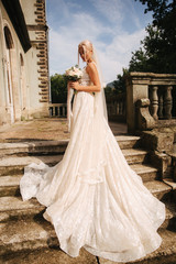 Obraz premium Gorgeous bride in wedding dress standing on the stairs by the palace. She hold a bouquet of flowers