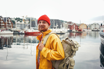 Side view of happy tourist with traveling rucksack standing among fishing boat on city pier. Alone...
