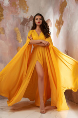 Young beautful caucasian woman with long wavy brunette hair in yellow flying dress posing against...
