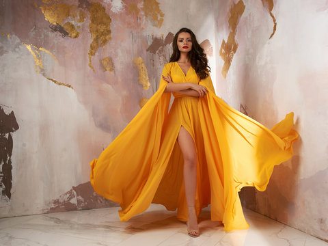Young beautful caucasian woman with long wavy brunette hair in yellow flying dress posing against wall. Vogue style fashion portrait