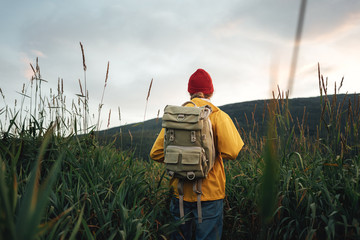 Hipster man traveler wearing backpack and yellow raincoat hiking among high grass. Young tourist...
