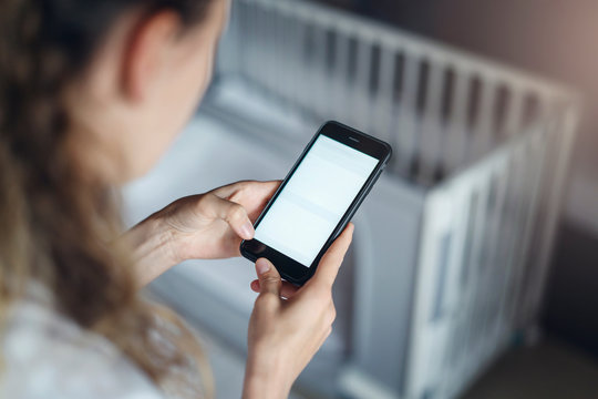Top view, close-up is smartphone in female hands. In background crib in soft focus. Mom businesswoman works remotely.