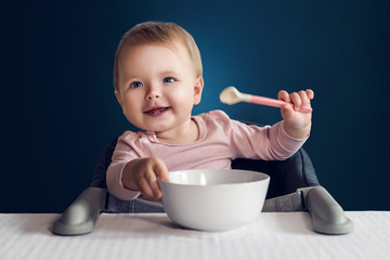 A nine-month-old smiling baby girl sits at a white table in a highchair and eats herself with a spoon from a bowl. Dark background