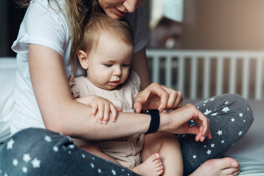 A nine-month-old baby girl with her mother is sitting on the bed and looking at the screen of a smart watch on her mother s hand.