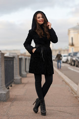 Portrait of young beautiful lady walking at street of city. Model wearing stylish black fur coat. Girl looking in camera. Female fashion concept.