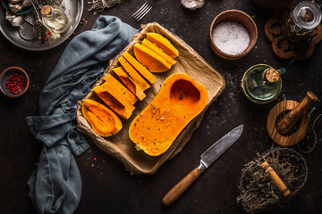 Butternut pumpkin halves on baking sheet. Cooking preparation on dark kitchen table background with herbs,spices, knife and utensils. Top view. Healthy seasonal food