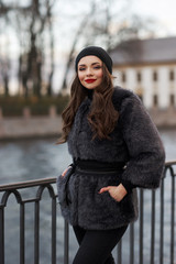 Pretty stylish woman with red lips and curls posing at river embankment in gray stylish fur coat and cashmere black hat