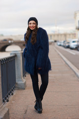 Happy joyful smiling young woman in blue lama fur coat and cashmere cap posing at city street. Day light portrait