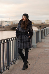 Beautiful lady in gray fur coat and cashmere cap at river embankment on a cold day. Fall winter fashion look.