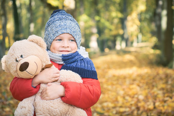 Little boy hugs his Teddy bear friend sitting on the ground in the autumn park.