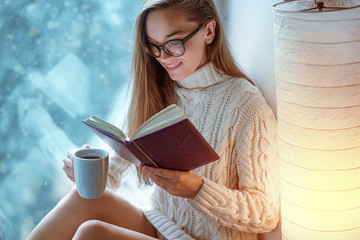 Obraz premium Portrait of comforting young happy cozy woman in glasses in knitted winter white warm sweater drinks a cup of hot cocoa during reading favorite book on a window sill by the window at home in winter