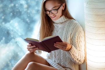 Happy attractive young cozy woman in glasses and knitted winter sweater enjoys reading a book on the window sill on the window at home in winter time.