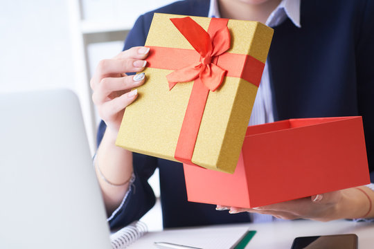 Christmas, any holiday. Office employee received a gift from her colleagues. Closeup woman hands holds gift box on office background.