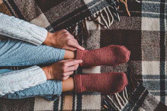 Female Feet In Knitted Winter Warm Socks And In Pajamas On Brown Checkered Plaid Blanket At Home In Cozy Winter Time. Top View