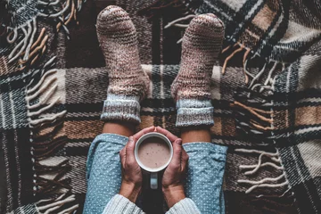Foto auf Acrylglas Cozy woman in knitted winter warm socks and in pajamas holding a cup of hot cocoa during resting on checkered plaid blanket at home in winter time. Cozy time and winter drinks. Top view © Goffkein
