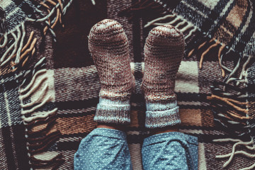Female feet in knitted winter warm socks and in pajamas on brown checkered plaid blanket at home in cozy winter time. Top view