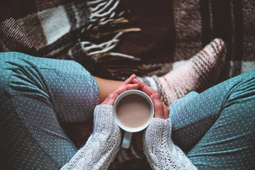 Foto op Aluminium Cozy woman in knitted winter warm socks and in pajamas holding a cup of hot cocoa during resting on checkered plaid blanket at home in winter time. Cozy time and winter drinks. Top view © Goffkein