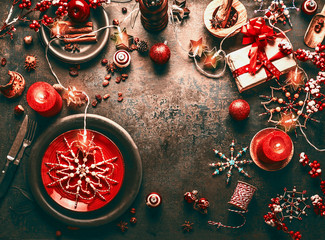 Christmas background with red festive dinner table setting, burning candles, gift box with red ribbon, snowflakes, cinnamon, anise, holiday decoration and fairy lights . Top view. Frame. Copy space