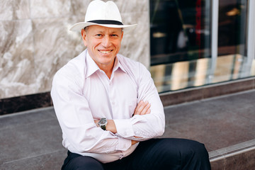 Portrait of  senior man in shirt and hat sitting on pavement folded hands . - Image