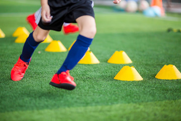 Kid soccer player putting red sport shoes to jogging between yellow marker cones on green artificial turf for soccer training of academy.