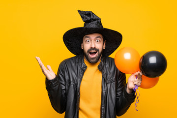 Man with witch hat holding black and orange air balloons for halloween party with shocked facial expression