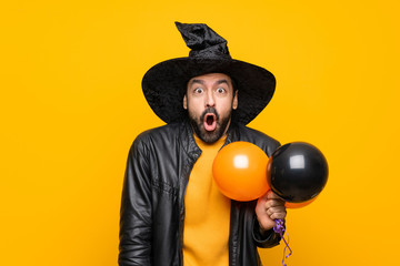Man with witch hat holding black and orange air balloons for halloween party with surprise facial expression