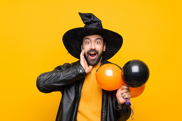 Man with witch hat holding black and orange air balloons for halloween party with surprise and shocked facial expression