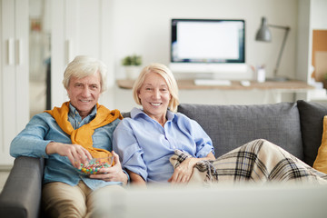 Portrait of modern senior couple watching TV together sitting on comfortable couch at home and smiling, copy space