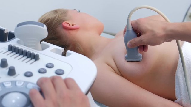 Young woman patient on ultrasound diagnostic of mammary glands on bust in clinic. Doctor runs ultrasound sensor over patient's mammary glands working on scanner panel. Diagnosis of internal organs.