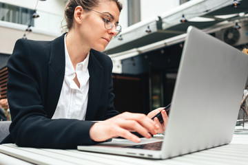 Fototapeta premium Young business woman wearing glasses at cafe using laptop and. Businesswoman sitting at coffee shop on terrace and working on computer