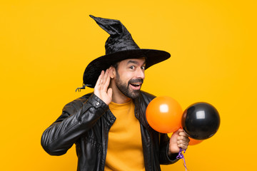 Man with witch hat holding black and orange air balloons for halloween party listening to something by putting hand on the ear