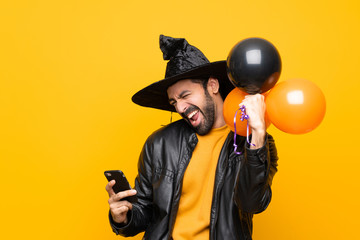 Man with witch hat holding black and orange air balloons for halloween party with phone in victory...