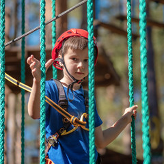 Boy enjoying activity in climbing adventure park at sunny summer day. Kid climbing in rope playground structure. Safe climbing with helmet insurance. Child in forest adventure park, extreme sport.