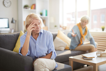 Portrait of senior couple fighting sitting on opposite sides of sofa at home, focus on crying wife in foreground, copy space