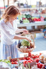 Happy mature woman buying fresh organic vegetables on a local farmer's marketplace. Holding a basket filled with various vegetables. Healthy food production and eating