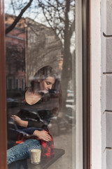 Obraz na płótnie Canvas Attractive girl with long brunette hair behind the glass, coffee shop window, concept of female emotions
