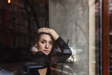 Fototapeta na wymiar Attractive girl with long brunette haibeside white flowers behind the glass, coffee shop window, concept of female emotions