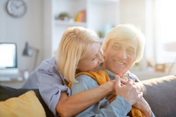 Warm-toned portrait of carefree senior couple embracing tenderly enjoying leisure time at home lit by sunlight, copy space