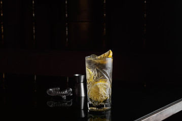 Alcoholic cocktail with lemon, gin and ice on black table near ice, jigger and strainer close up
