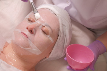 Masked woman on anti-aging procedure in a beauty salon. Facial skin cleansing in a cosmetology center. Beautician applies a white brush to a transparent gel on the girl's forehead.